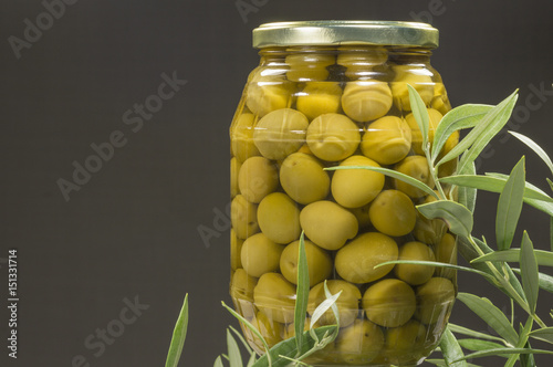 Un recipiente de cristal con aceitunas en conserva, hojas de olivo junto al recipiente