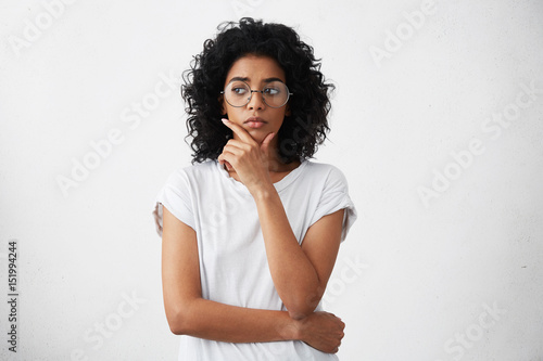 Portrait of beautiful casually dressed young woman in round glasses having doubtful expression, looking away in indecisiveness, holding her chin, trying to find best solution. Body language