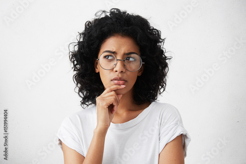 Isolated portrait of stylish young mixed race woman with dark shaggy hair touching her chin and looking sideways with doubtful and sceptical expression, suspecting her boyfriend of lying to her