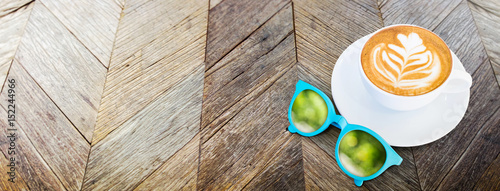 Hot coffee cup with sunglasses and reflection of green tree bokeh on rustic old wood plank table top view,Mock up banner for adding your text or design