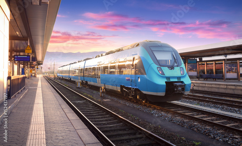 Piękny widok z nowoczesnym pociągiem dużej prędkości na stacji kolejowej i kolorowe niebo z chmurami o zachodzie słońca w Europie. Krajobraz przemysłowy z niebieskim pociągiem na peronie kolejowym. Tło kolejowe