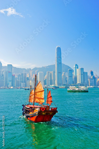 View of Hong Kong skyline with a red Chinese sailboat passing on the Victoria Harbor in a sunny day.