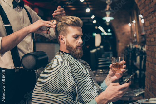 Chill out at the barber shop. Side view of handsome young red bearded man drinking scotch and browsing at his pda, while getting a haircut