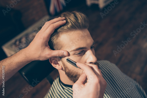 Close up of a hairdresser`s work for an attractive young blond man at the barber shop. He is doing styling of his beard, shaving it with straight razor