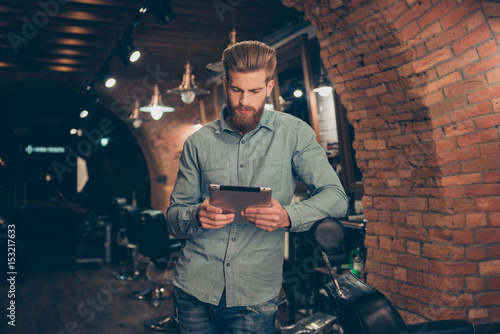 Casual men`s life. Perfect! Harsh stylish red bearded man in a barber shop is browsing on his tablet, serious and concentrated, waiting to get a haircut