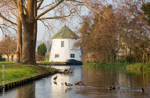 Provincial spring-winter-autumn Dutch landscape with ice-covered canal,blue boat, white rural house and feeding ducks.Reflection of trees and house in still water of small Netherlands canal.