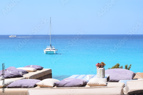 Luxuriöse blaue Yacht ankert im türkisfarbenen Wasser vor der Küste von Formentera, im Vordergrund Bänke und Kissen am Ufer, im Hintergrund ein Motorboot