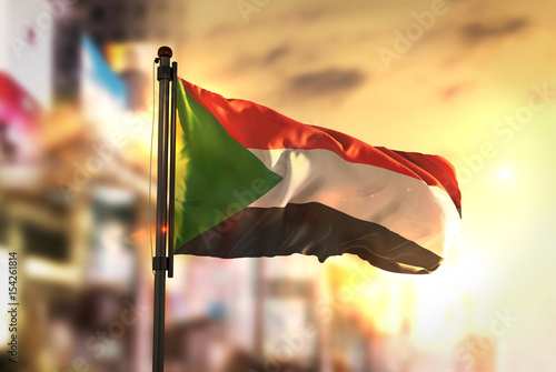 Sudan Flag Against City Blurred Background At Sunrise Backlight