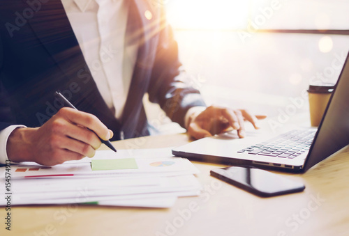 Businessman working at sunny office on laptop.Man pointing notebook keyboard and holding pen hand.Blurred background.Papaer documents on the table.Sunlight effects.
