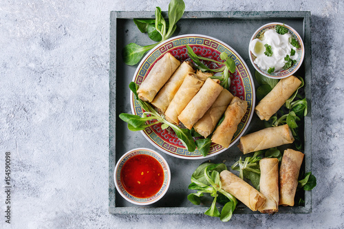 Fried spring rolls with red and white sauces, served in china plate on square wood tray with fresh green salad over gray blue texture background. Flat lay, space. Asian food