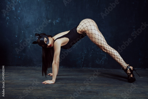 Beautiful sexy cat-girl is standing in plank on hands in a black slip, Leather cat mask, in black pantyhose, in black high hills on dark background. BDSM concept