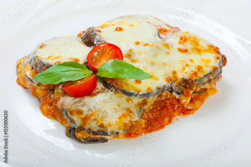 Baked eggplant with cheese, tomatoes and spices on a white plate. A dish of eggplant is on a wooden table.