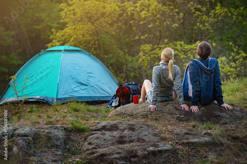 couple of hikers with backpacks resting near the tent in the warm sunshine at sunset. Travel, vacation, holidays and adventure concept. Forest Mountain landscape background
