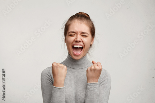 Cheering happy young Caucasian female with pure skin and beautiful appearance clenching fists being excited of success. Joyful woman screaming with happiness to achieve her goals. Good luck concept