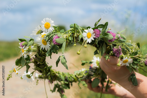 Hands making flower chain of chamomile and clover on sunny spring meadow with dirt road in the background.