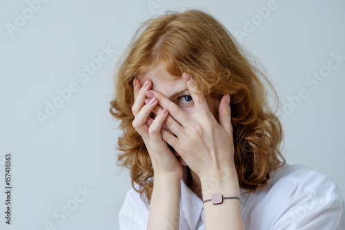 A red-haired girl with curls in embarrassment covers her face with her hands and looks through her fingers. Social phobia of the younger generation.