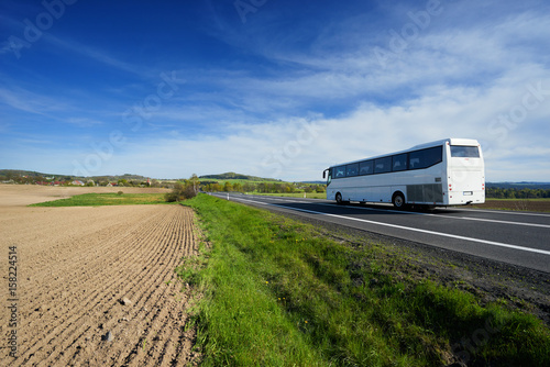 The white bus traveling on the road around a sown field in a rural landscape with a village in the background