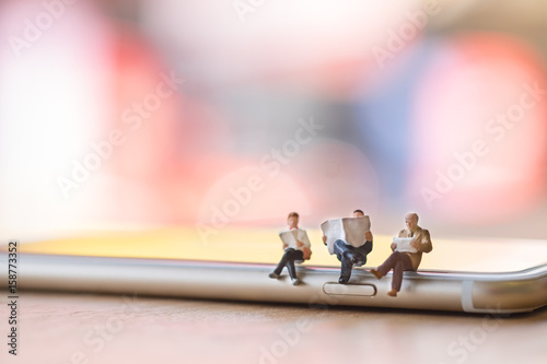 Group of miniature figures sitting and reading a book and newspaper on smart phone on wooden table.