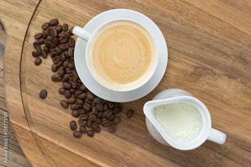 Coffee concept: Top view of white porcelain cup filled with delicious frothy coffee, jug of milk and fresh roasted coffee beans on rustic wooden background.