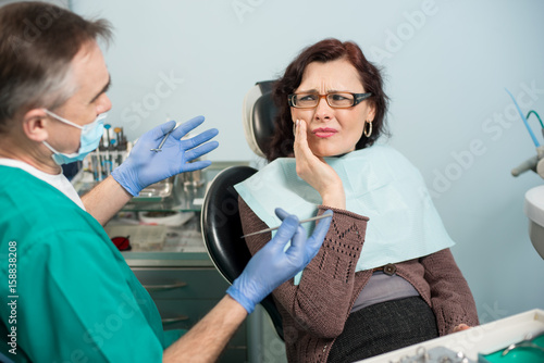 Woman feeling toothache, touching cheek with hand at dental clinic. Senior dentist trying to help. Dental care and health concept