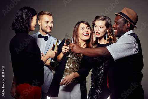 Group of well-dressed colleagues clinking champagne flutes together while having joyful office party, pretty woman whispering something in her friends ear