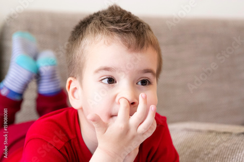 Kid scratching a finger in his nose. Itching boy lying on his stomach with head in hands and legs bent at the knees while watching TV.