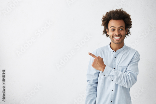 Horizontal portrait of dark-skinned handsome man having broad smile wearing formal white shirt posing against white background pointing with index finger at white copy space for your advertisment