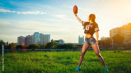 Young athletic girl in short denim shorts and a sports shirt posing against a background in a city park on a sunset background and holding a soccer ball
