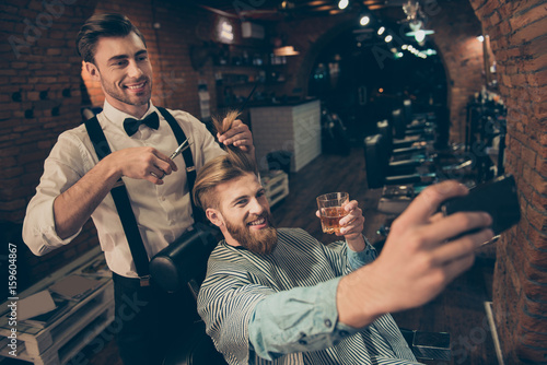 Smile! Cheerful handsome stylish red bearded guy is taking selfie photo at barber shop, classy dressed smiling stylist is making him a brand new haircut and posing also
