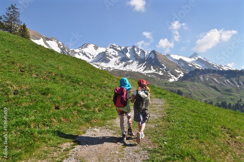 Zwei Kinder wandern Hände haltend auf Wanderweg in den Schweizer Bergen auf Wanderweg. Grüne Wiese und Schneeberge im Hintergrund