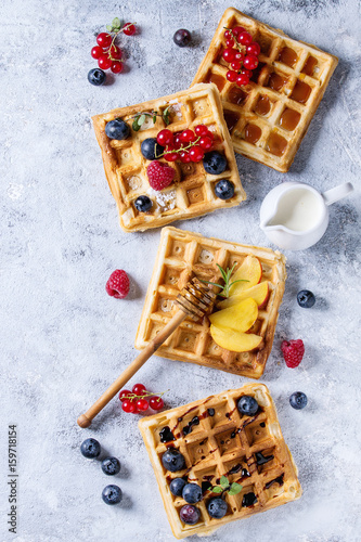Homemade square belgian waffles with fresh ripe berries blueberry, raspberry, red currant, peach served with caramel, balsamic sauce, honey, whipped cream over gray background. Top view with space