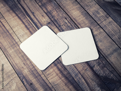 Two blank square beer coasters on wood table background.