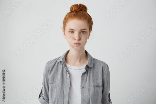 Portrait of grumpy woman with freckled face, blue eyes, ginger hair and eyebrows blowing her cheeks being dissatisfied with her results. Pretty red-haired girl making grimace having upset look