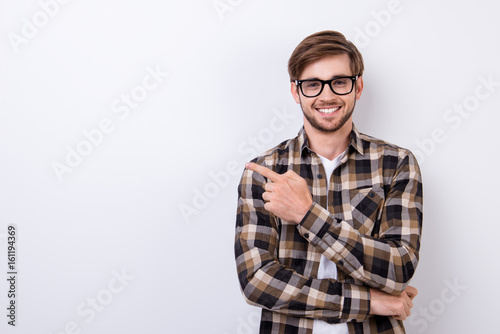 Smiling young nerdy bearded stylish student is standing on pure background in glasses and casual outfit, pointing on the copyspace