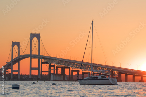 Sunset over Newport Bridge in Newport, Rhode Island. There is a sailboat moored in front of the Newport Pell Bridge
