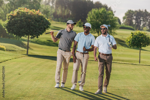 Confident smiling men in caps and sunglasses holding golf clubs and walking on lawn