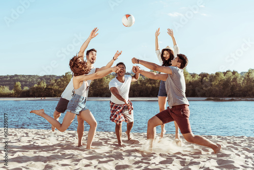 young smiling friends playing beach volleyball on riverside at daytime