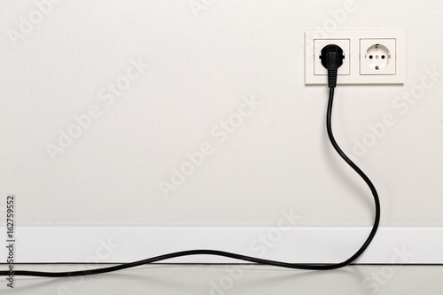 Black power cord cable plugged into european wall outlet on white plaster wall