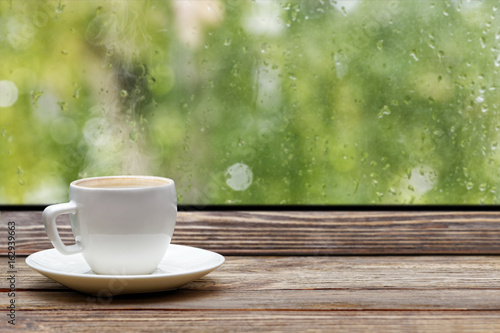 White steaming cup of hot coffee on vintage wooden windowsill or table against window with raindrops and foliage on blurred background. Shallow focus.