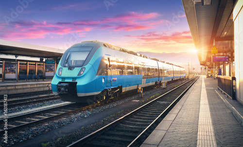 Nowoczesny wysokiej prędkości kolejki na stacji kolejowej i kolorowe niebo z chmurami o zachodzie słońca w Europie. Przemysłowy krajobraz z niebieskim pociągiem pasażerskim na peronie kolejowym. Tło kolejowe