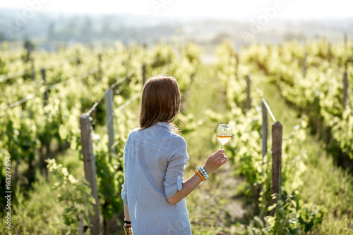 Young woman with wine glass standing on the beautiful vineyard during the sunset