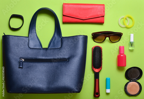 Women's fashion accessories on a green background. Leather bag, red purse, comb, sunglasses, cosmetics, perfume, smart watch. What does a woman have in her bag? Top view. Flat lay.