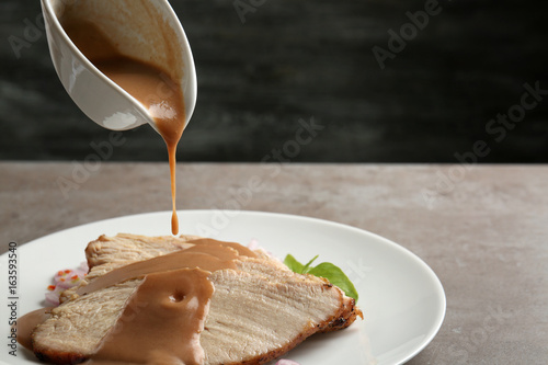 Pouring tasty turkey gravy onto sliced meat on plate against dark background