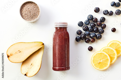 Healthy smoothie drink with fresh fruit including pear, blueberries, lemon and chia seeds arranged around a bottle of blended juice on a white background
