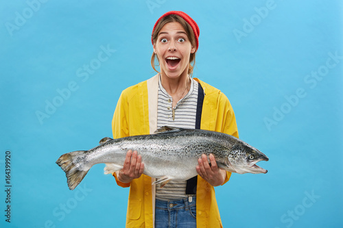 Wife of fisherman holding huge fish having surprised expression looking with bugged eyes and jaw dropped not believing her eyes rejoicing successful catch. Happy shocked fisherwoman with trout