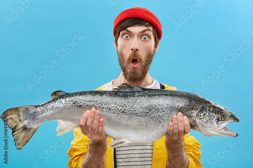 Indoor shot of astonished bearded fisherman dressed casually holding huge fish looking at camera with bugged eyes and jaw dropped being shocked to catch such big trout or salmon. Surprisment