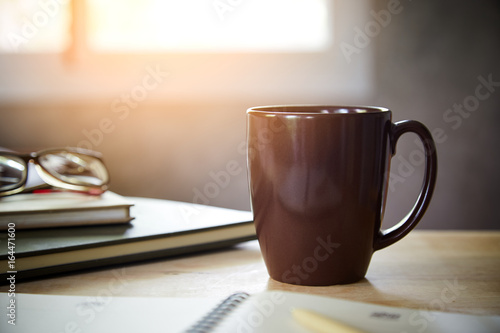 Morning coffee on the desk with sunlight from window, Coffee mug on the table with the blurred of notebook and glasses