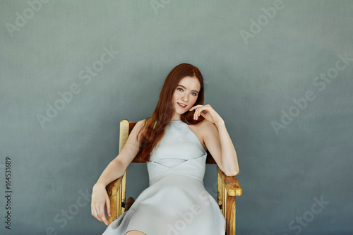 Studio portrait of stunning fashionable young European female model with long ginger hair, dressed in cocktail dress sitting on wooden chair against blank wall with copy space for your content