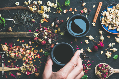 Chinese black tea in black stoneware cups, man's hand holding one cup and wooden spoons with dry herbs, flower buds and leaves over black wooden background, top view, horizontal composition