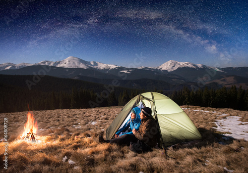 Romantic couple tourists having a rest in the camping at night, sitting near campfire and green tent under beautiful night sky full of stars and milky way. On the background snow-covered mountains